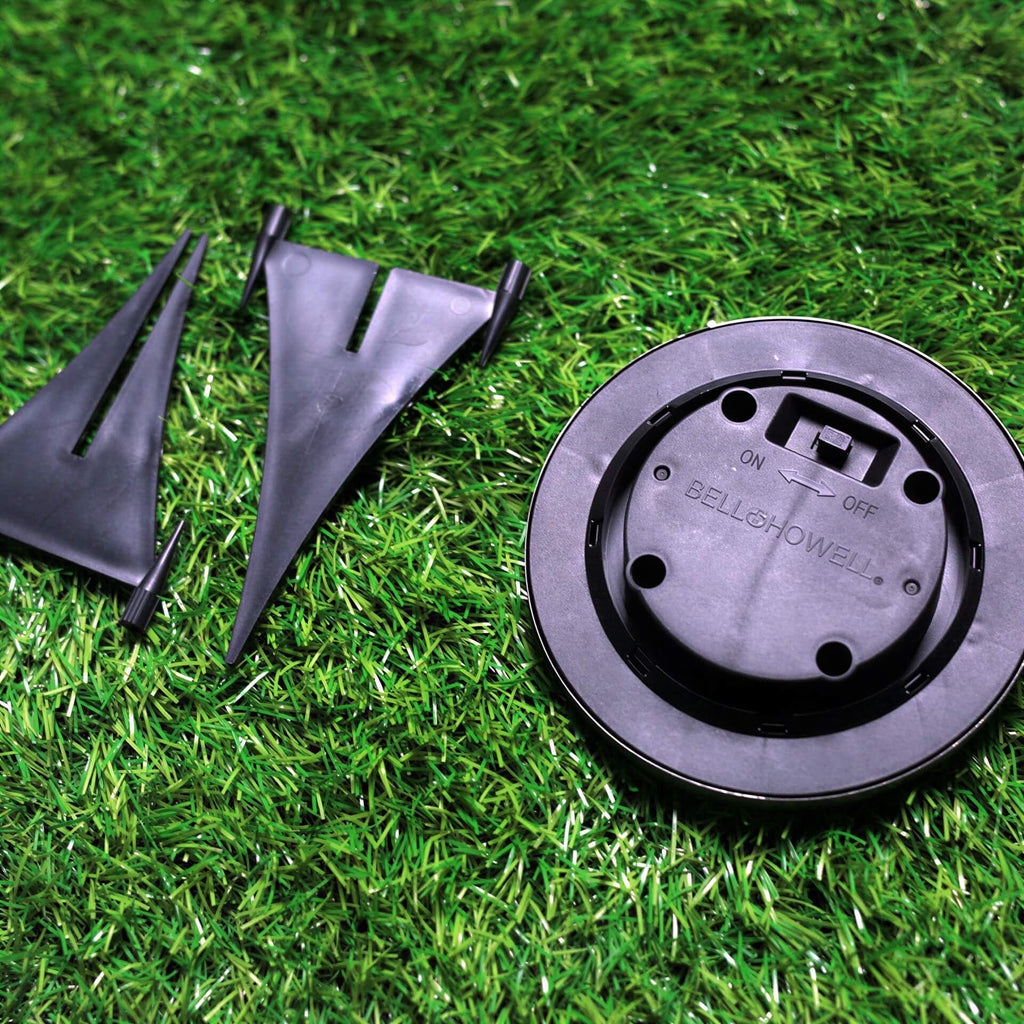 Round black plastic solar light base and two pointed stakes on green grass. Label reads BELL+HOWELL and ON OFF switch.
