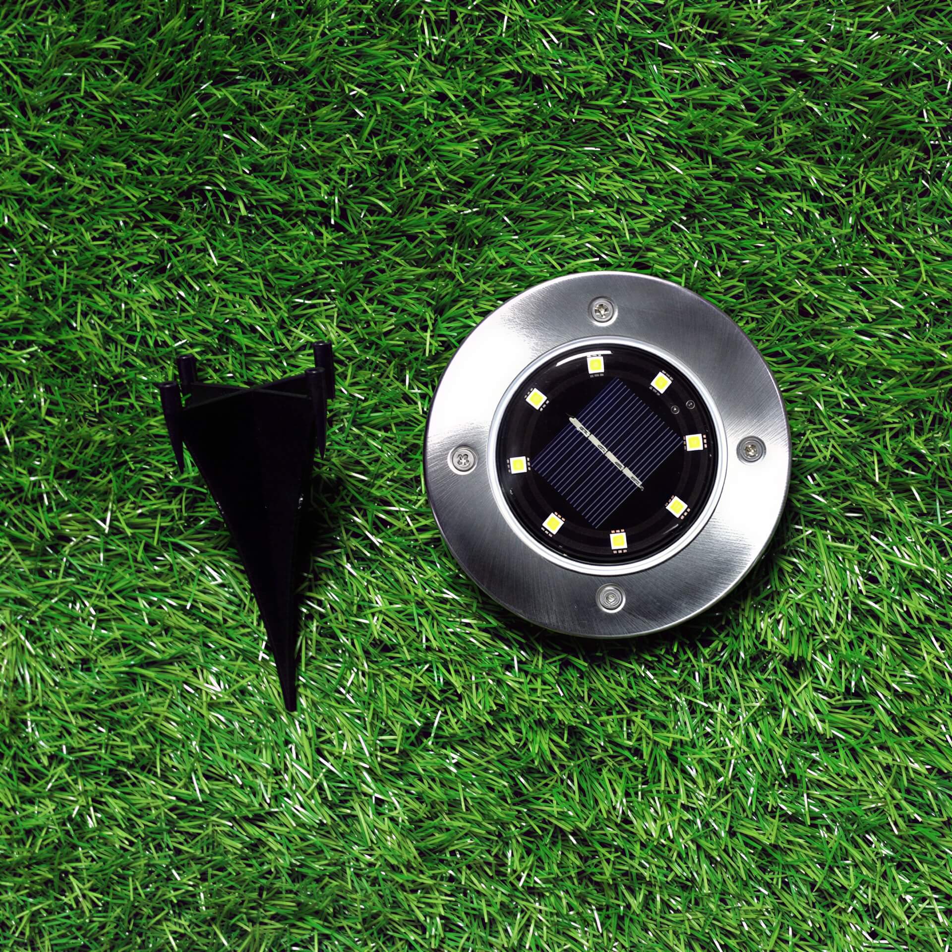Round metal solar ground light with eight LEDs and a black plastic stake on green grass.