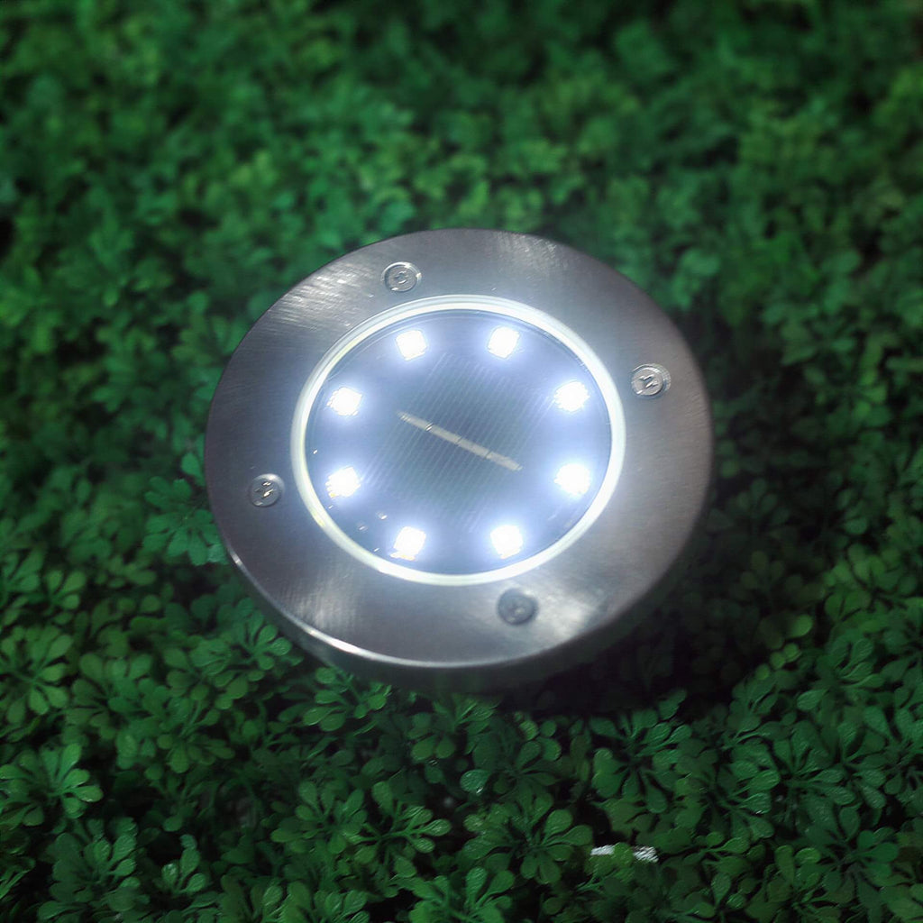 One round metal solar ground light with eight glowing LEDs and a center solar panel on green foliage.